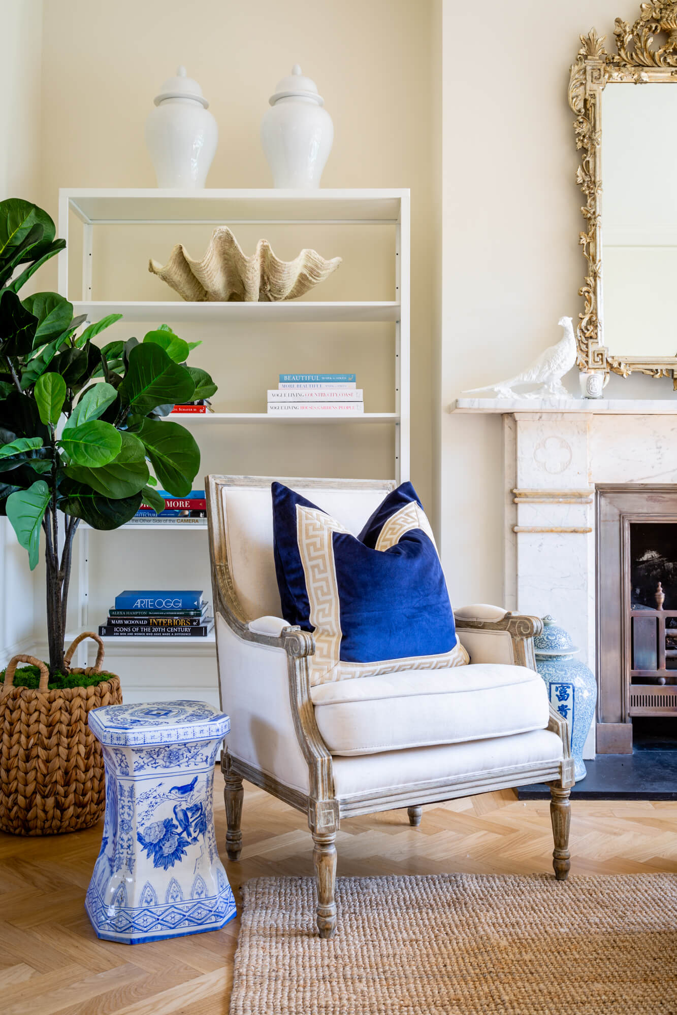 A French linen armchair surrounded by blue and white ginger jars, blue and white stool, decorative items and a large French gilt mirror on top of a fireplace