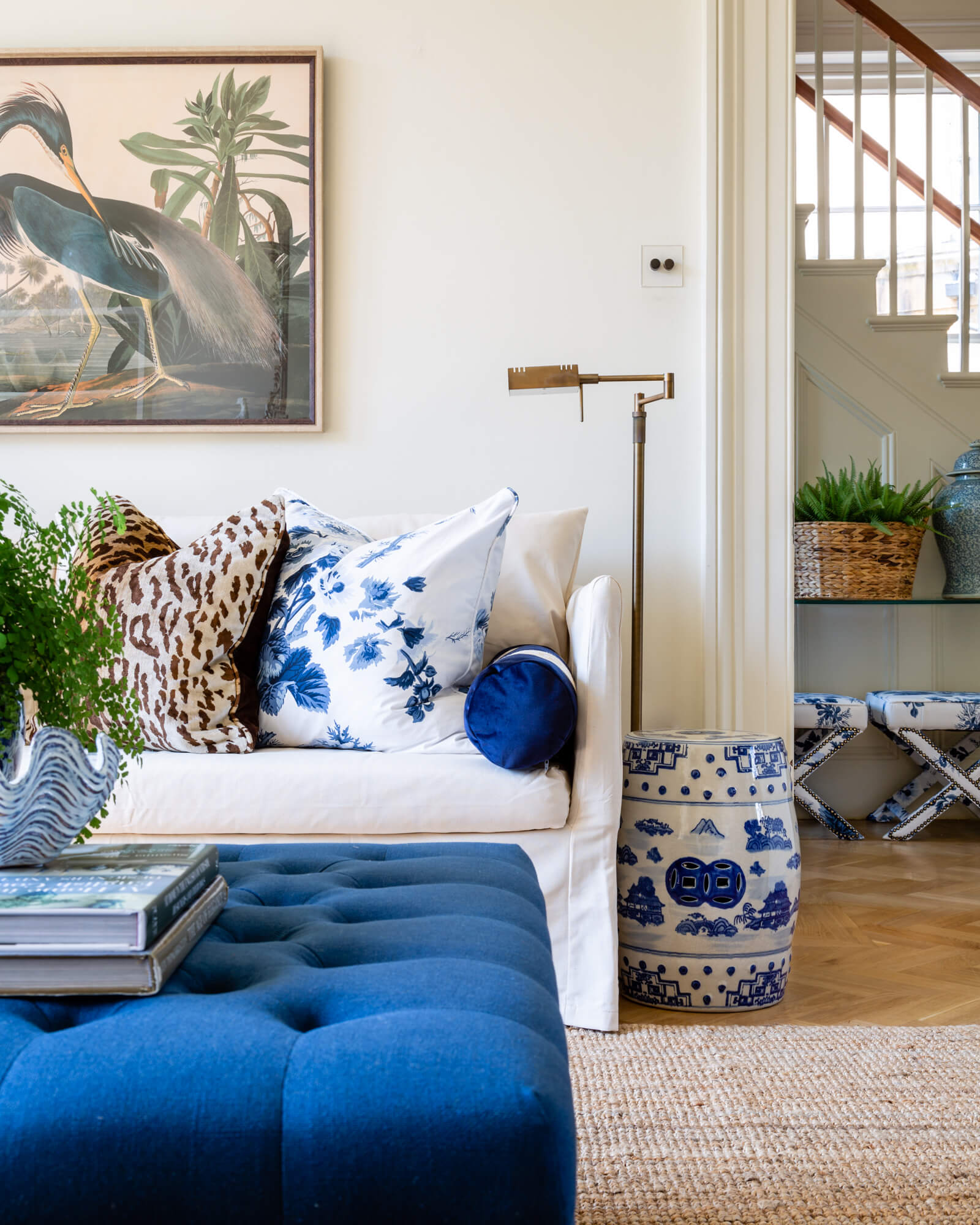A living room with a white linen sofa, leopard cushions, a blue linen footstool, blue and white stools, and a heron bird painting on the wall