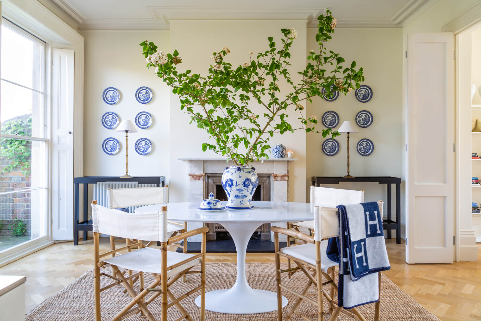 A dining room with a blue and white ginger jar on top of an oval white Saarinen table surrounded by director’s chairs on a sisal rug.