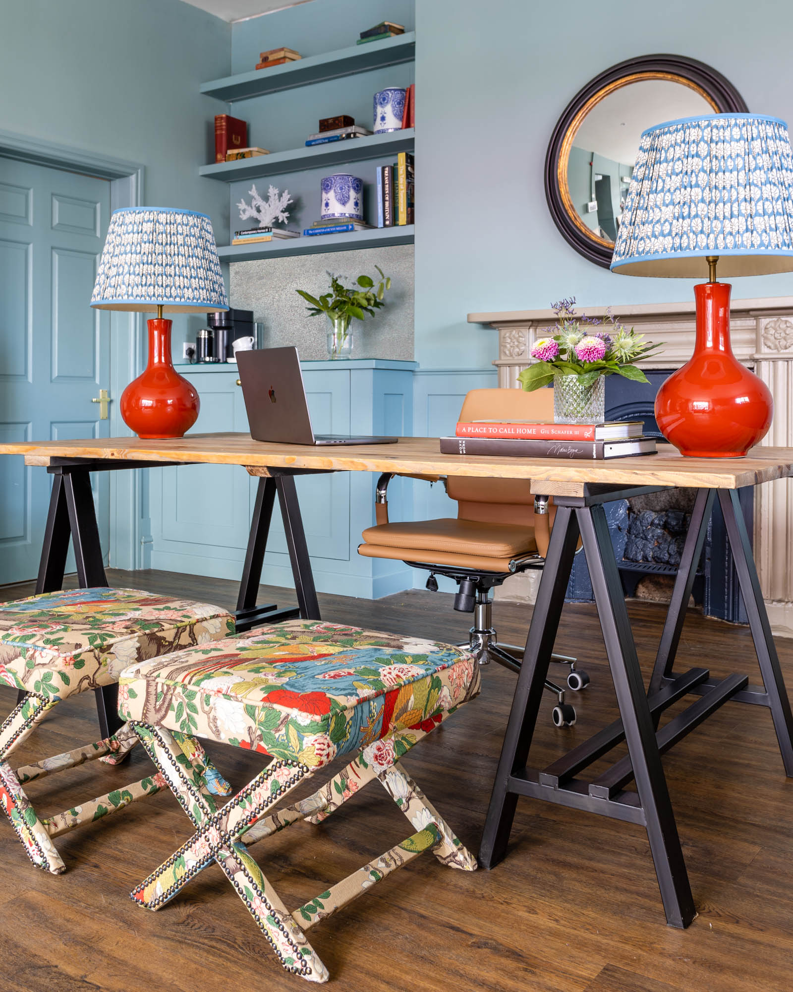 A straight on shot of the desk area in a boutique real estate agency. A trestle desk sits at the centre, with red Pooky lamps on either side, and GP & J Baker’s Rockbird Signature botanical print on the x-frame stools below. The walls are painted in Farrow & Ball’s Oval Room Blue, and behind the desk is featured a round convex mirror. To the left is a coffee station with shelving above, featuring vintage books, ginger jars and decor items chosen by the client.