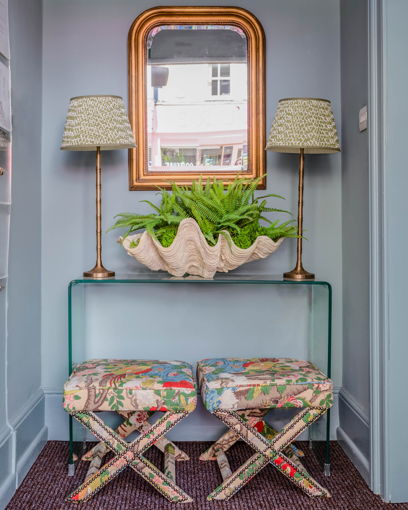 The entryway of a boutique real estate agency, features Farrow & Ball’s Oval Room Blue on the walls, and a glass console table, on which is placed two Pooky lamps with green shades and a large clamshell overflowing with fern fronds. Below this sit two bespoke x-frame stools upholstered in GP & J Baker’s Rockbird Signature botanical print.